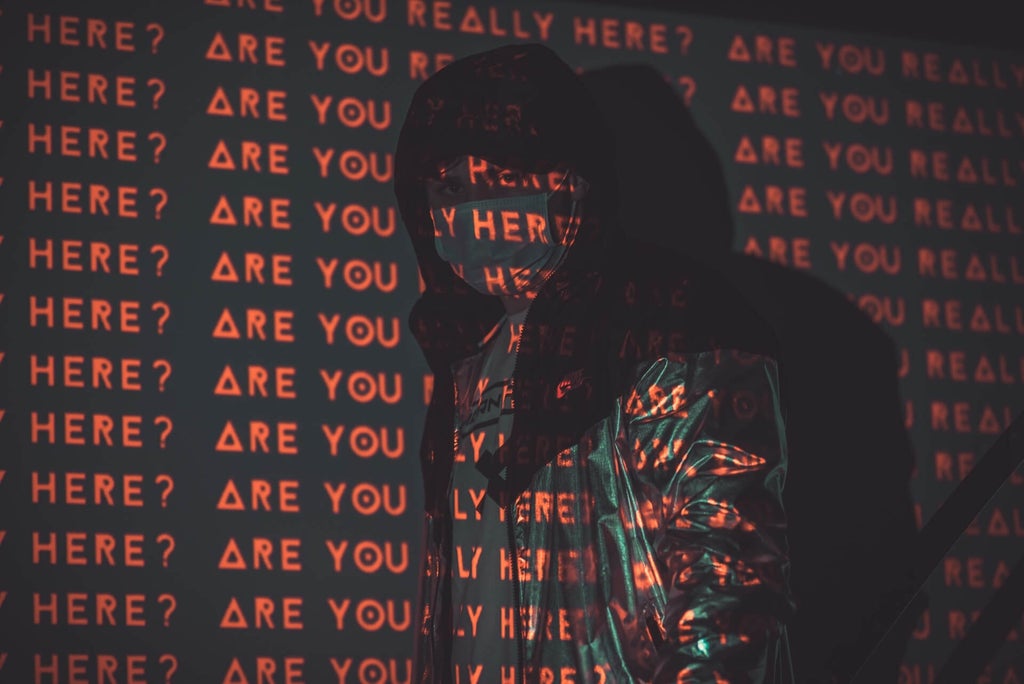 Person stands in dark with the phrase "Are you here?" repeatedly projected across them in red light.